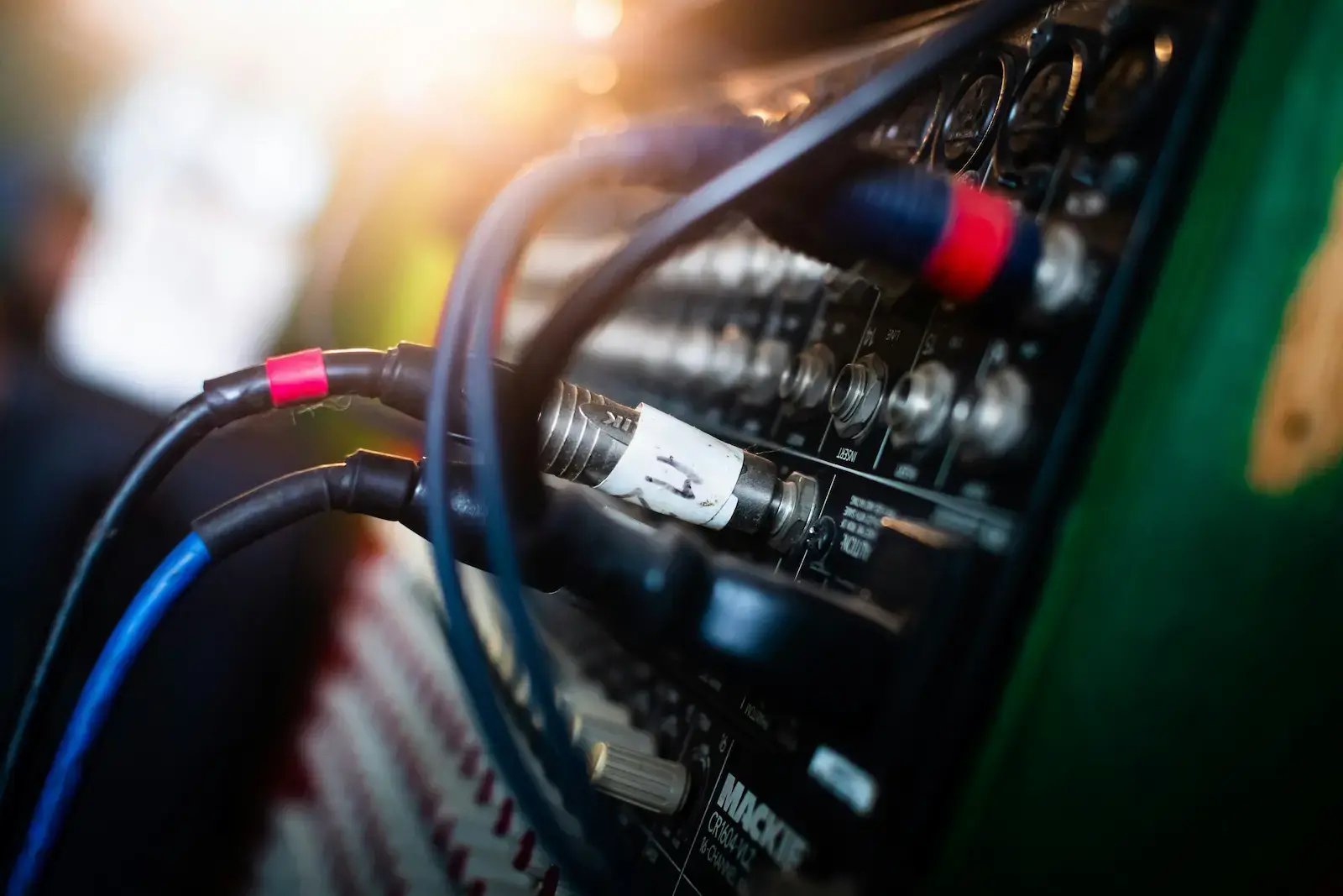 close-up of audio cables plugged into a mixing board with labeled inputs and a sun flare in the background