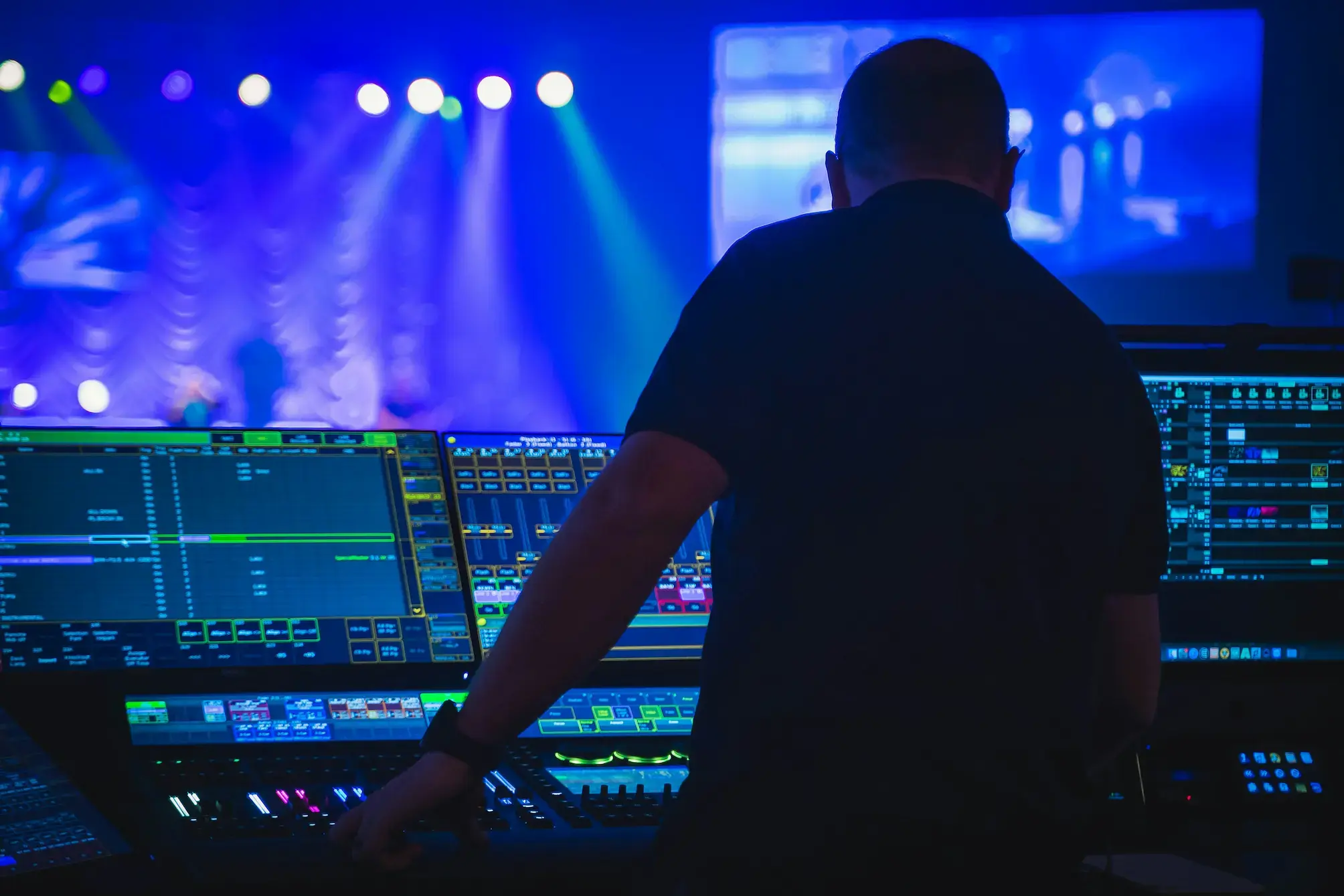 audio technician standing at large digital mixing console with glowing screens during a live stage event with blue lighting